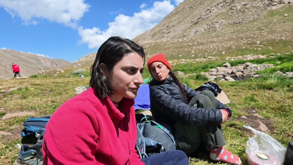 Two young women sitting on the grass with their rucksacks below a stony slope, one in a pink fleece and one in a dark padded jacket and a red knitted hat.