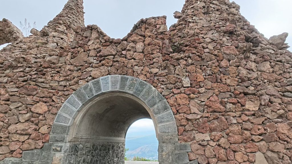 A ruined wall of rough reddish porous stone with a neat grey stone archway, through which hazy mountains and a valley are visible.