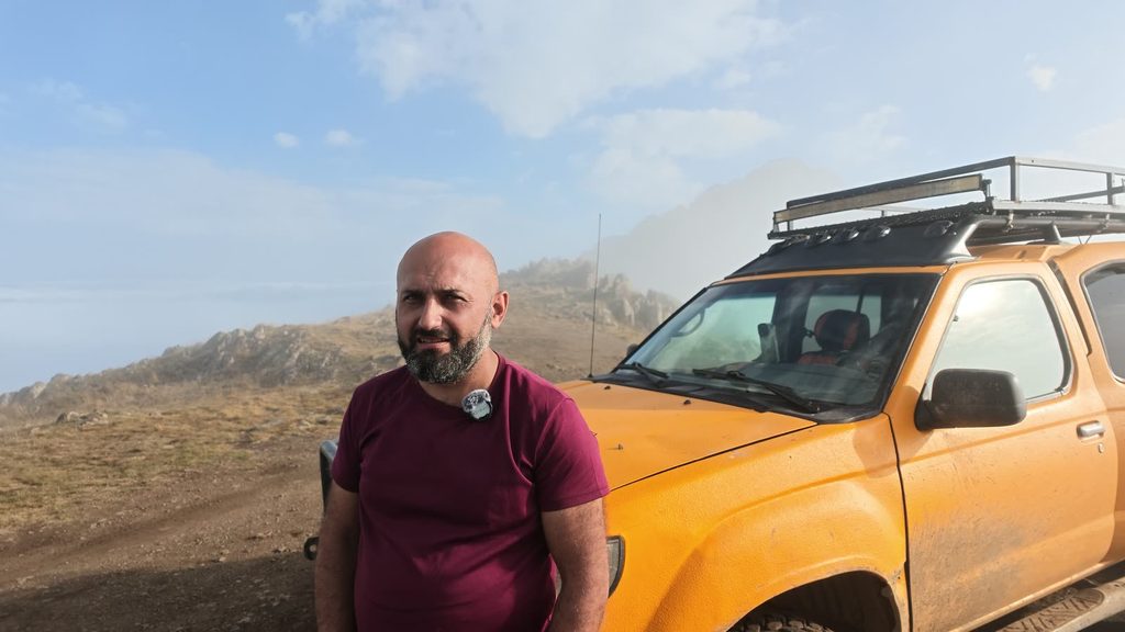 Harut, a bald man with a grey-flecked beard in a maroon T-shirt, standing beside the yellow four-by-four on the plateau.