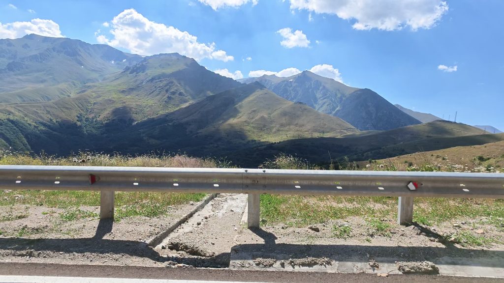 Green mountains and a valley under a blue sky with white clouds, seen over a metal crash barrier at the roadside.