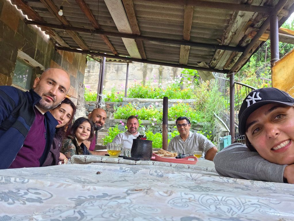 Seven people around a table under a covered veranda, with glasses, a teapot and plates on a patterned cloth and a garden beyond.