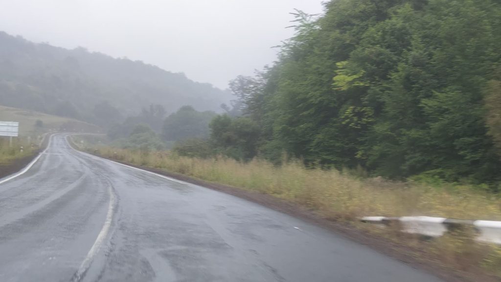 A wet mountain road curving into thick mist between forested slopes, taken from a moving car.