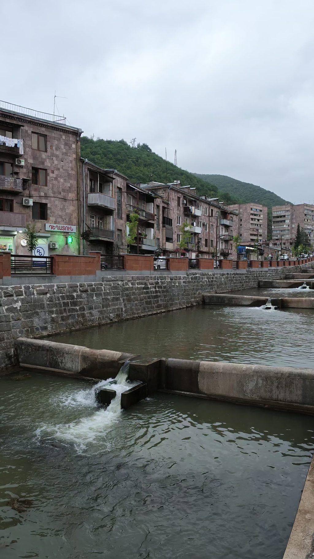A river running through a stone channel and stepping down over a series of low concrete weirs, beside a row of pink stone apartment blocks under a green hill.