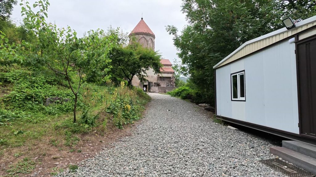 A gravel path leading past a white prefabricated cabin towards the stone church of Vahanavank, with its conical dome, among green trees.