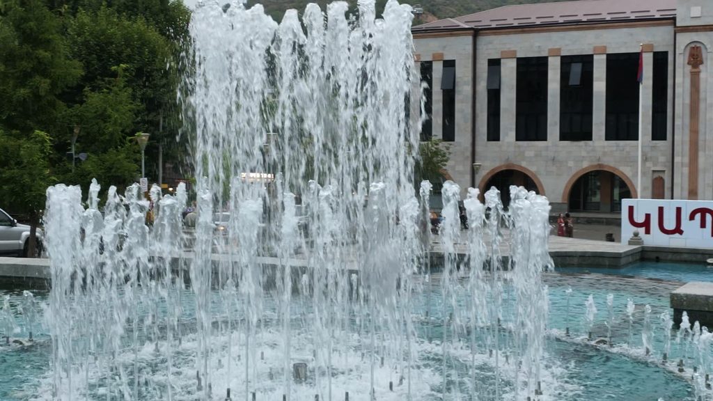 A fountain with dozens of tall water jets in a turquoise basin, in front of a pale stone civic building with arched entrances and an Armenian flag.