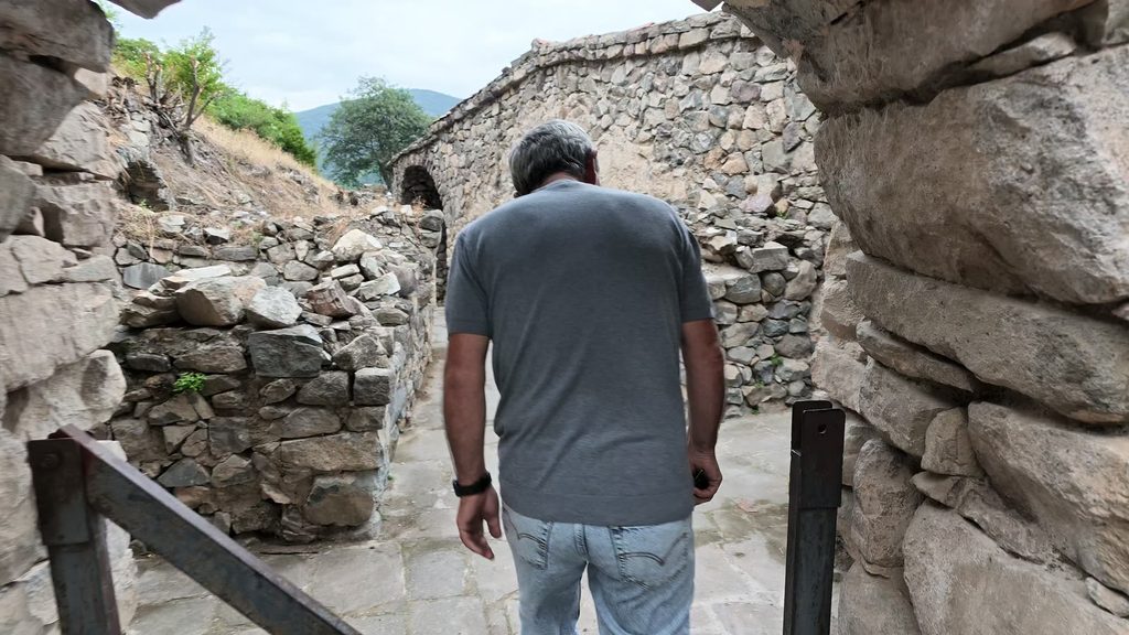Aram, seen from behind in a grey T-shirt, walking along a stone passage at Halidzor fortress towards a vaulted stone structure.
