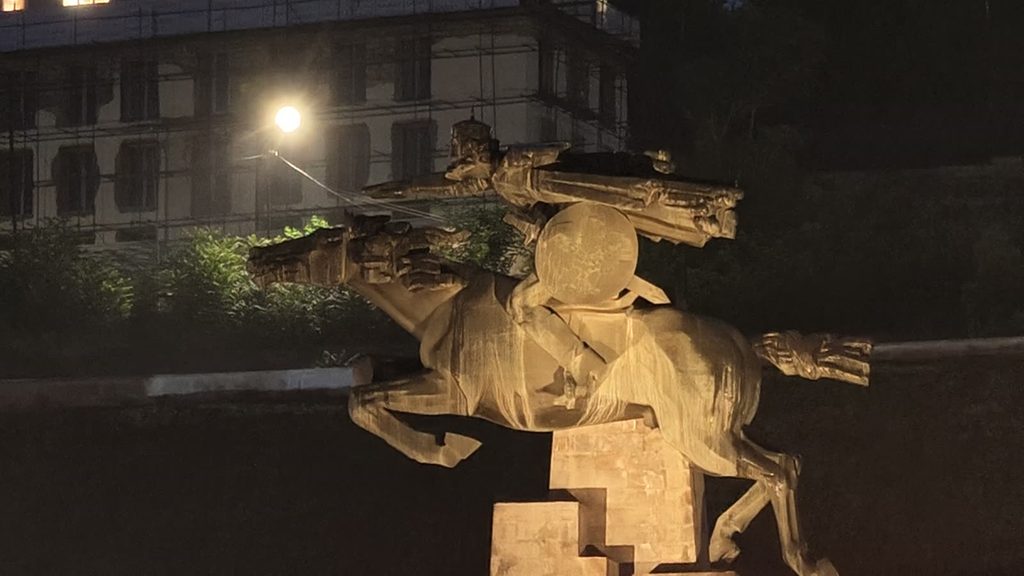 A floodlit statue of a rider with a round shield on a rearing horse, on a stepped stone plinth at night, with a scaffolded building behind.