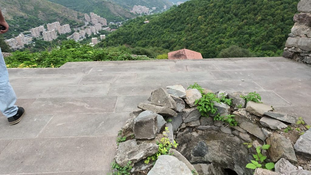 The apartment blocks of Kapan in a green valley, seen from a stone terrace at Halidzor fortress with a ruined round wall in the foreground.