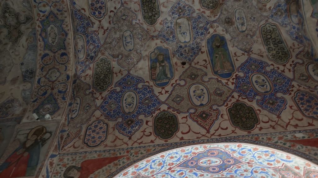 A church vault painted with blue, red and olive-green ornament on a cream ground, with medallions holding figures of saints.