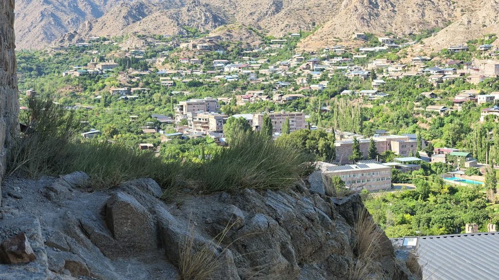Meghri from above: houses and apartment blocks among green trees in a valley ringed by bare brown mountains, seen past a rocky ledge.