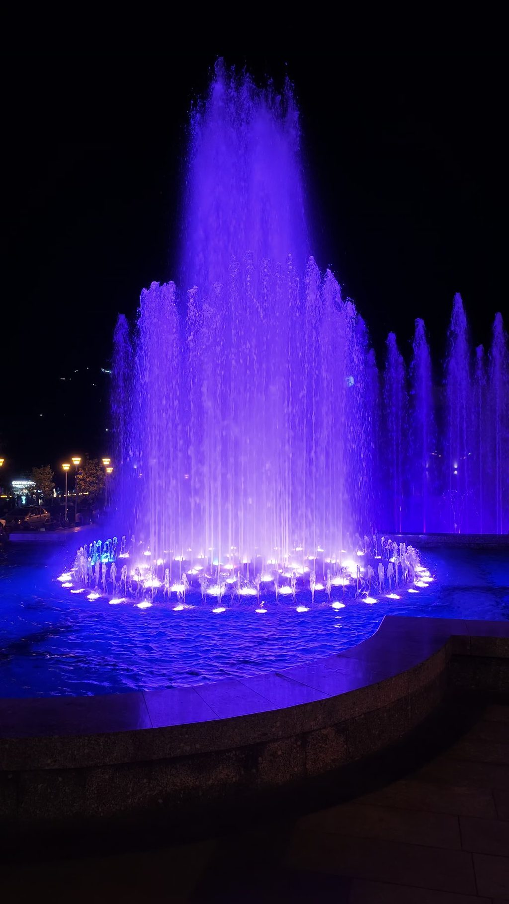 A fountain lit purple and blue at night, its jets rising from a round basin.