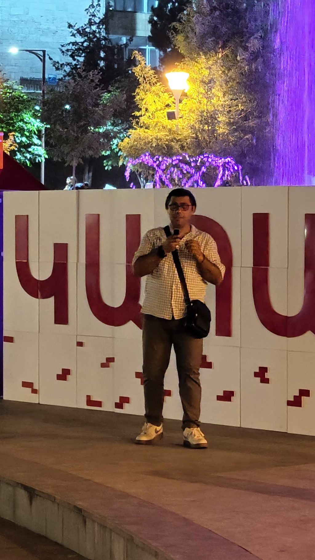 Garo in glasses and a checked shirt, a shoulder bag at his side, speaking into a phone in front of large dark-red letters on white panels spelling Kapan in Armenian, at night.