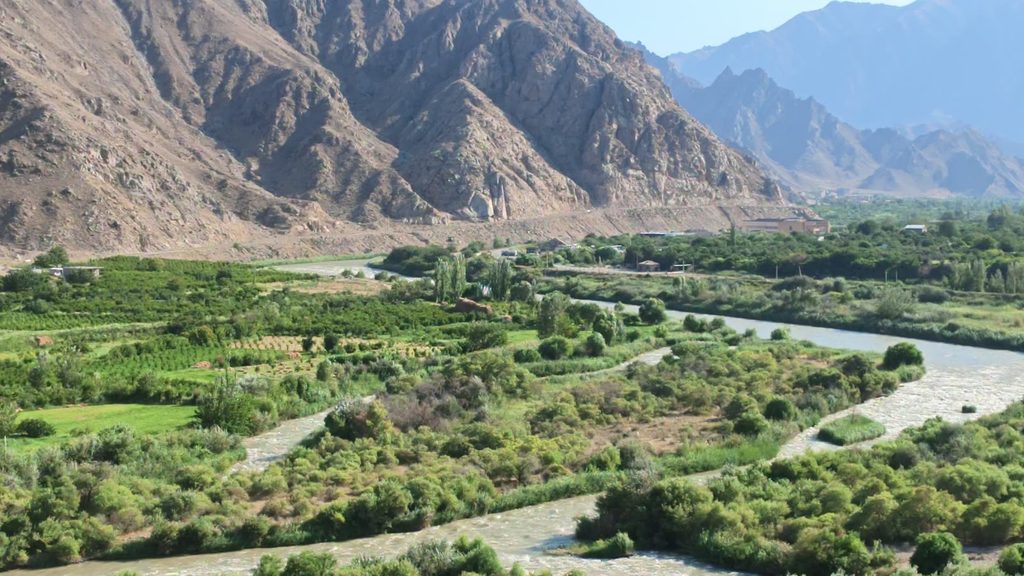 The grey river Araks running between green banks and orchards, with steep brown mountains rising on the far side under a blue sky.