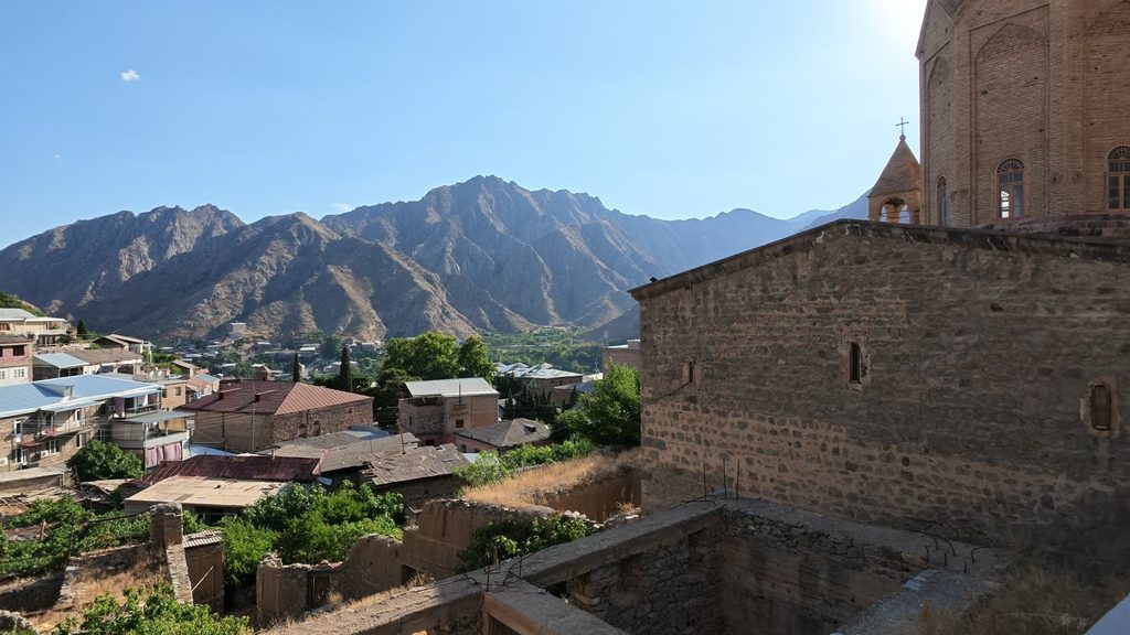 The brick church of Surb Astvatsatsin and its small belfry at the right edge, above an old stone wall and the metal roofs of Meghri's old houses, with bare mountains beyond the town in the evening sun.