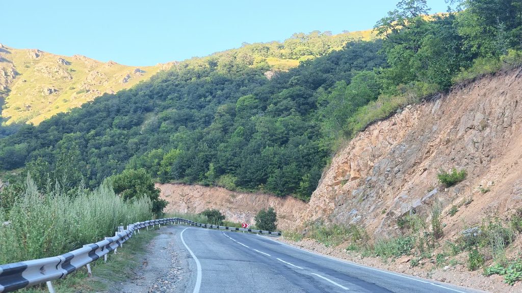 A winding mountain road in evening sunlight, with a crash barrier, a cut rock face and a forested slope above.