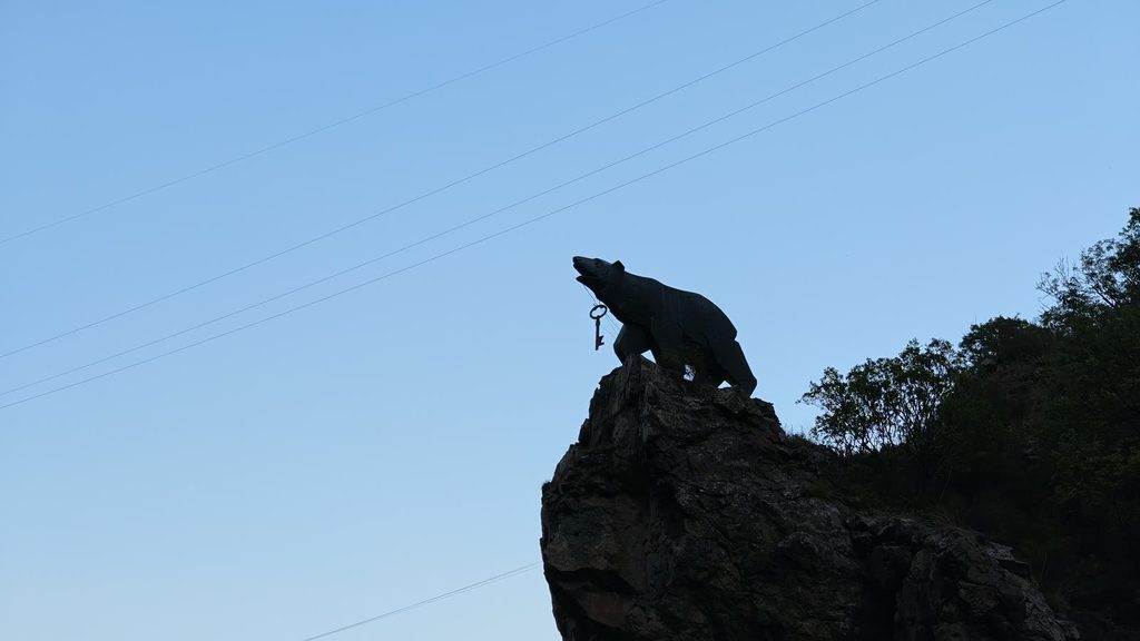 The dark silhouette of a bear statue holding a key in its mouth, on a rock against a pale blue sky.