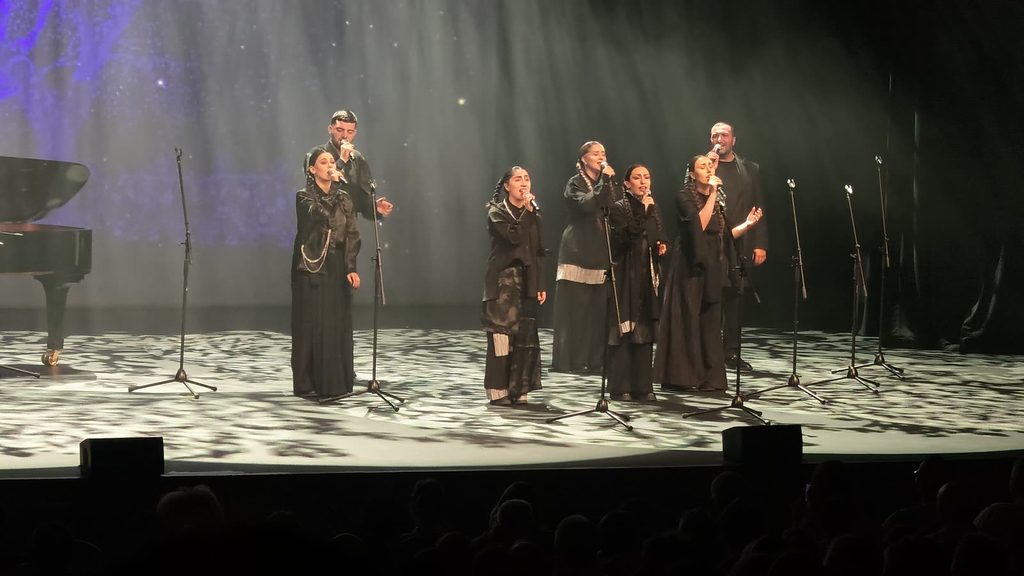 Seven singers in black costumes stand among microphone stands on a theatre stage, lit by dappled white light beside a grand piano, with the audience in silhouette below.