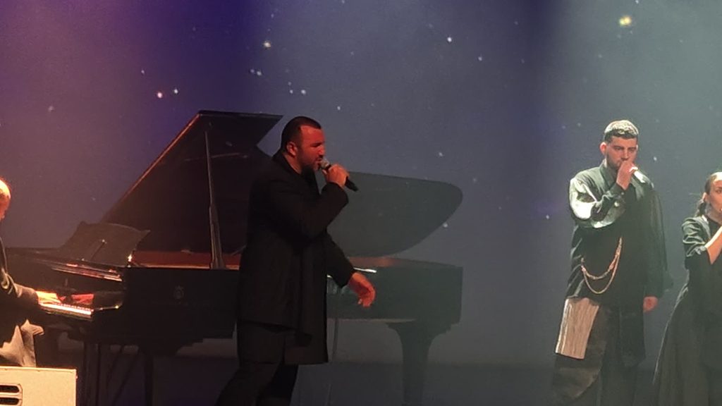 A singer in a long black coat sings in front of an open grand piano while the pianist plays at the left edge and two more singers perform on the right.