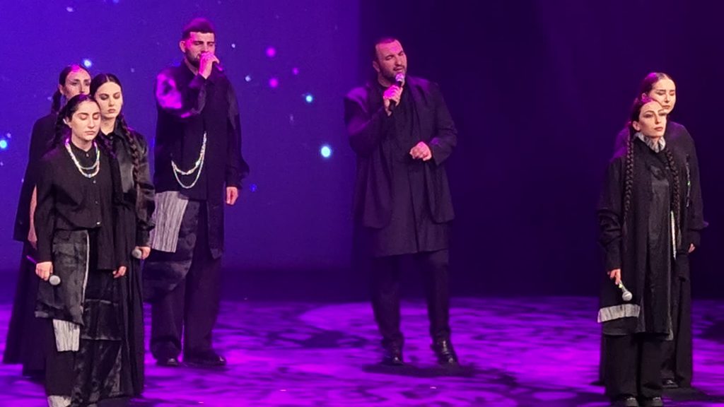 Two men sing at the centre of a stage bathed in purple light while women in black costumes and long braids stand on either side with eyes closed.