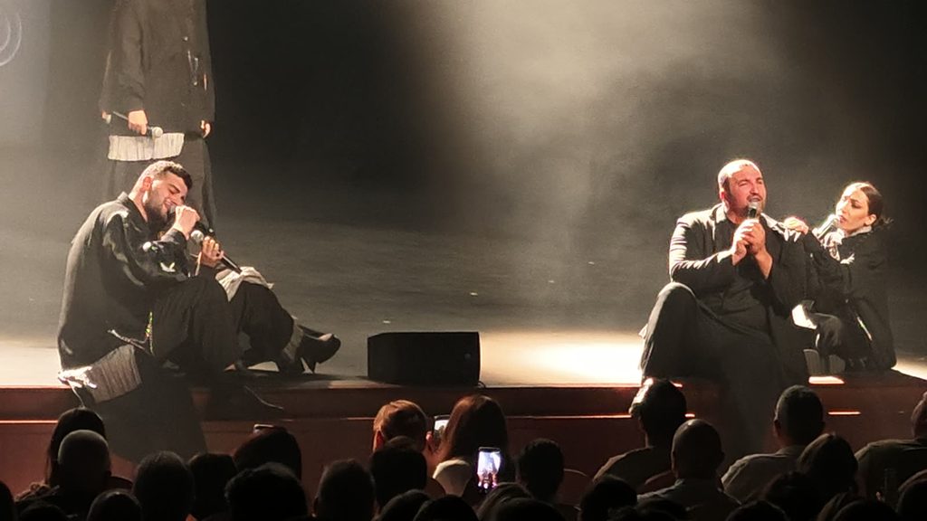 Singers sit at the front edge of the stage singing into microphones just above the front row, where audience members hold up phones to film them.