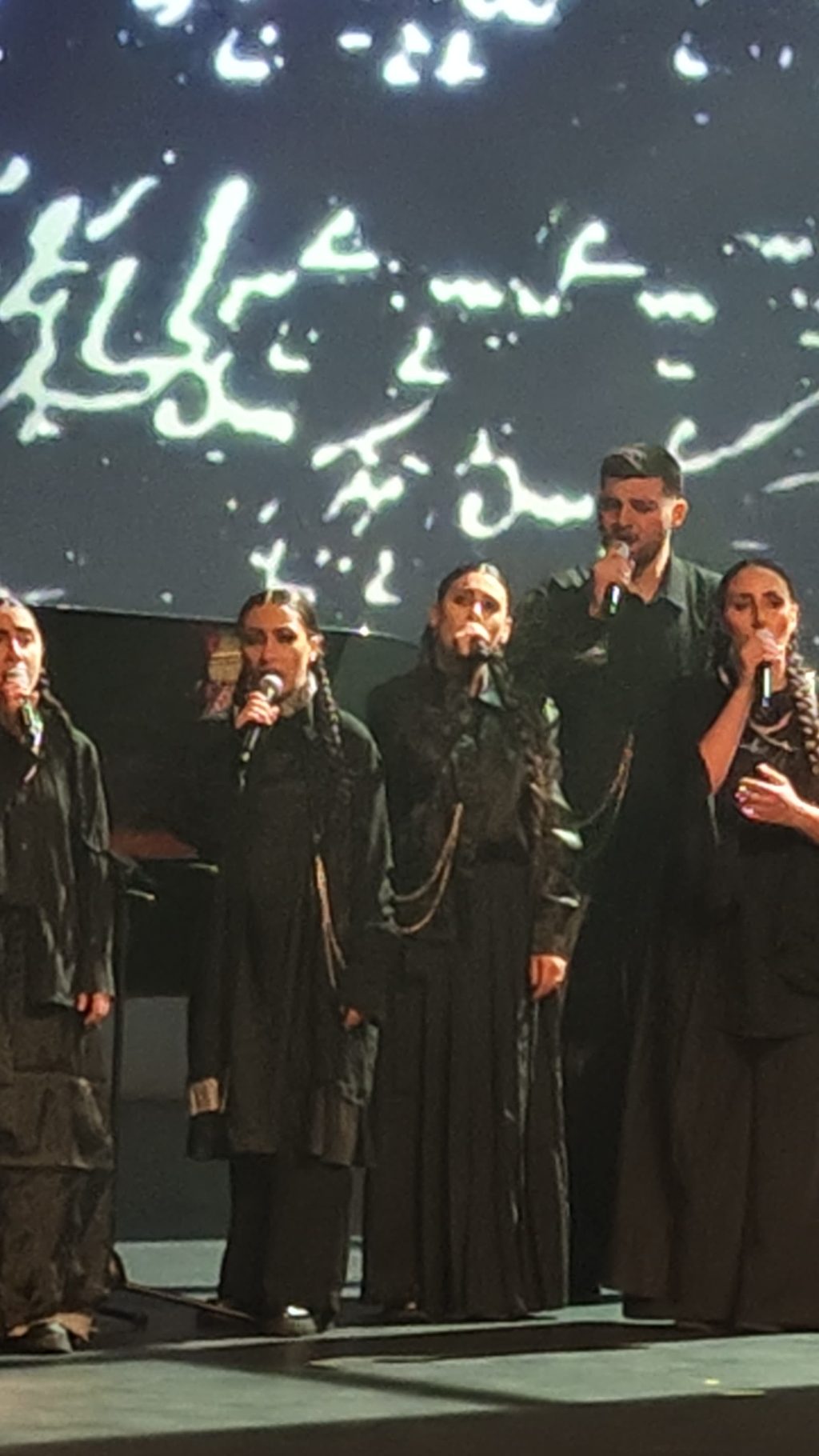 Singers in black costumes sing into microphones beneath a large screen showing white handwritten notation projected behind them.