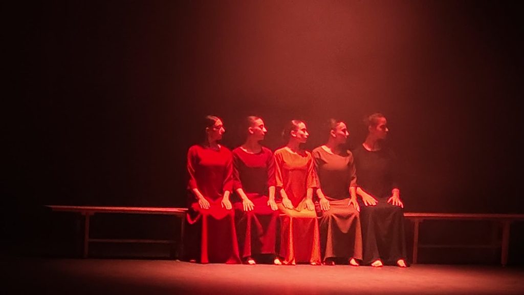 Five barefoot dancers in long plain dresses sit side by side on a wooden bench under red stage light, all turning their heads to the same side.