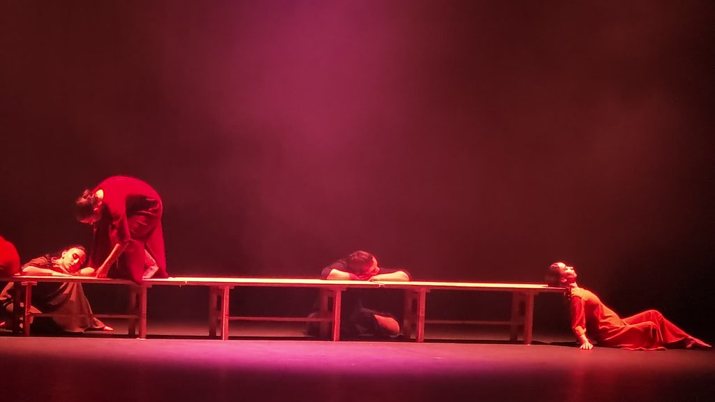 Dancers in long dresses slump and lean against a row of wooden benches under magenta stage light, one reclining on the floor with her head tipped back against the bench.