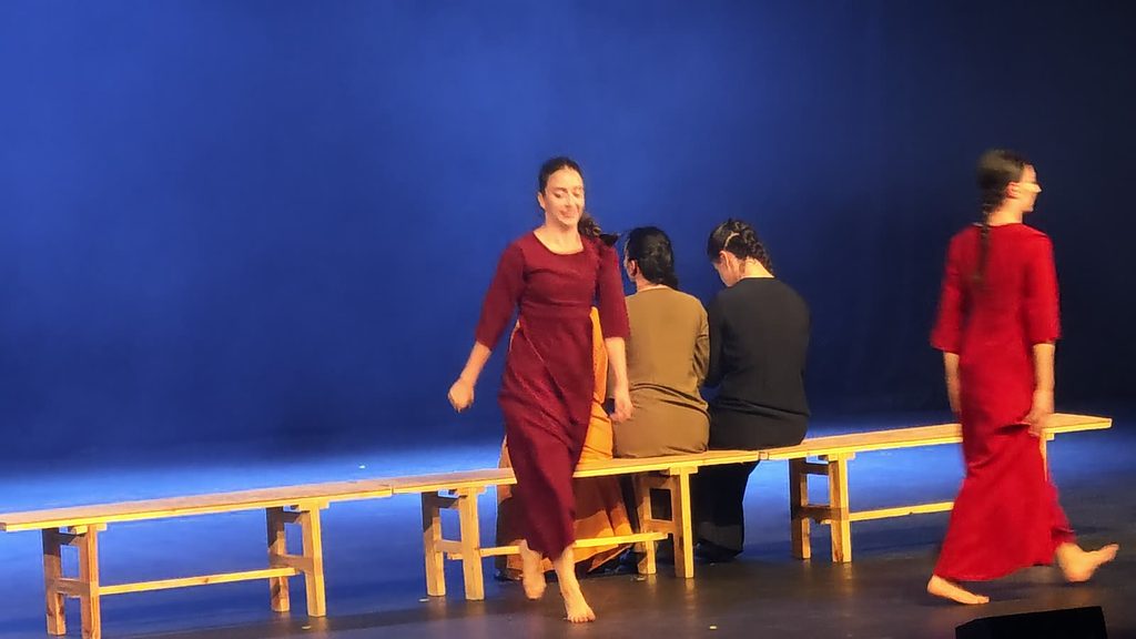 Against a deep blue backdrop, a smiling dancer in maroon walks barefoot past a bench where dancers in orange, olive and black sit facing away, while a dancer in red crosses at the right.