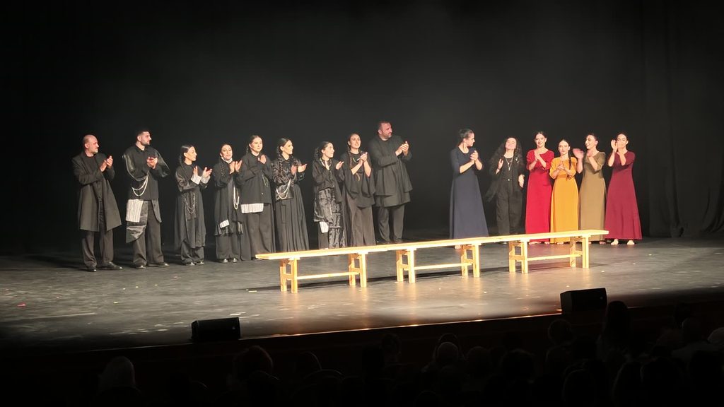 At the curtain call, a row of singers in black and dancers in coloured dresses stand clapping across a dark stage behind three wooden benches, with the audience in silhouette below.