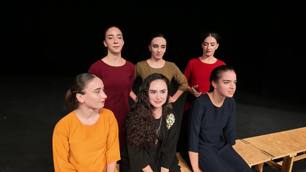 A smiling woman with long curly hair, dressed in black with a floral brooch, sits among five young dancers in orange, maroon, olive, red and navy dresses.