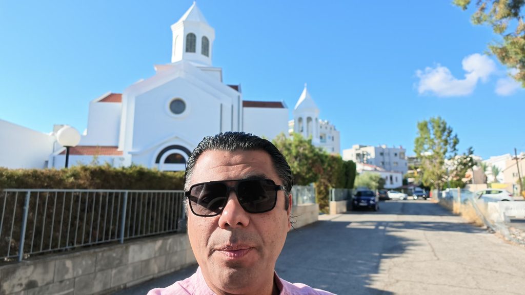 Garo Kotchounian in sunglasses in front of Sourp Asdvadzadzin, the white Armenian cathedral of Nicosia, under a blue sky.