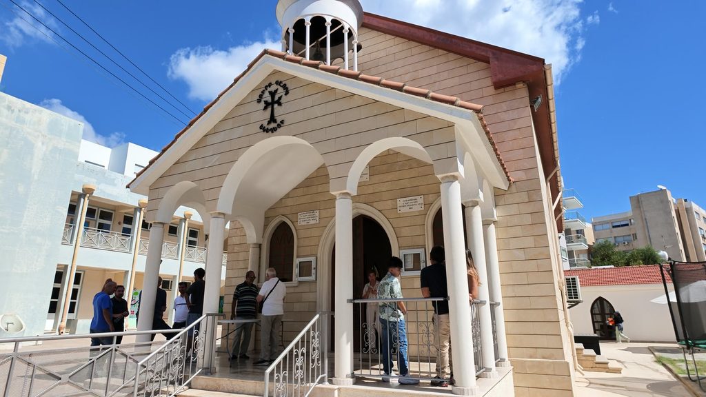 The small Armenian church of Sourp Kevork in Limassol, clad in pale stone, with a porch of white arches and a white bell cupola, people standing on the porch and the school building to the left.