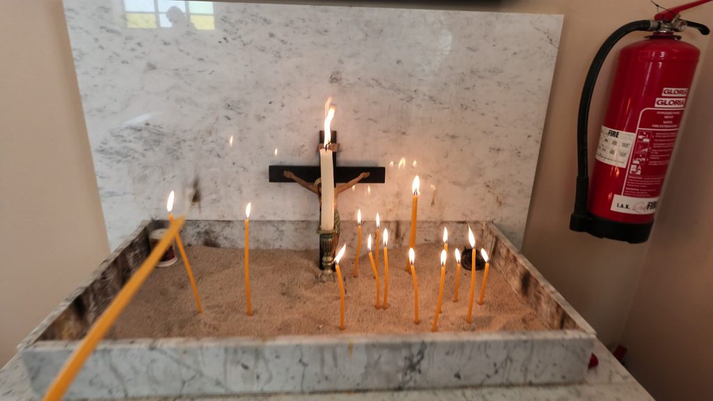 Thin yellow candles burning in a tray of sand before a small crucifix, against a marble wall.