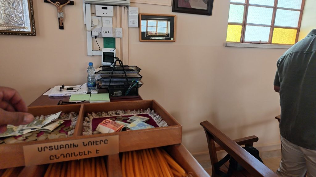 A wooden candle box with trays of coins and banknotes, above a drawer of yellow candles, with a card in Armenian capitals reading ՍՐՏԱԲՈՒԽ ՆՈՒԷՐԸ ԸՆԴՈՒՆԵԼԻ Է.