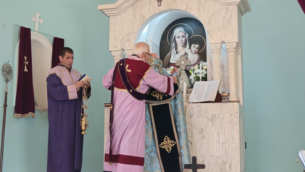 Seen from behind, Archbishop Gomidas Ohanian in blue and gold and a server in rose and burgundy stand at a marble altar beneath a painting of the Virgin and Child, while a second server in purple holds a censer.