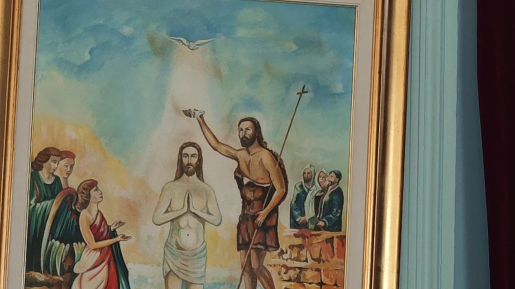 A painting of the Baptism of Christ in a gilded frame on a pale green wall: John the Baptist pours water over Christ as a dove descends, with angels and onlookers at either side.