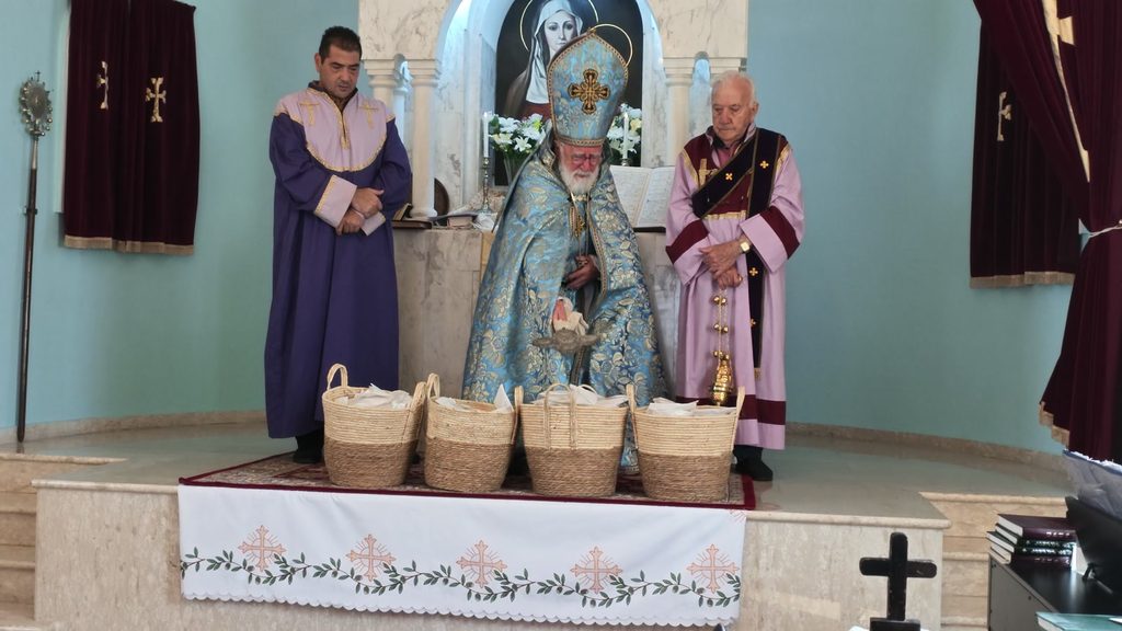 Archbishop Gomidas Ohanian in blue and gold bends over four straw baskets of white-wrapped portions on the altar step, a hand cross in his hands, between two servers in purple and in rose and burgundy.