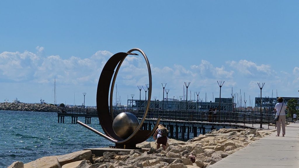 A steel sculpture of a ring around a polished sphere on the rocks of the Limassol seafront, beside a long wooden pier, with yacht masts and the marina behind.