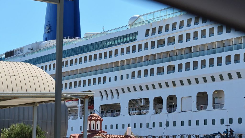 The long white side of the cruise ship Aurora berthed at Limassol port, with the red-tiled belfry of a small chapel in the foreground.