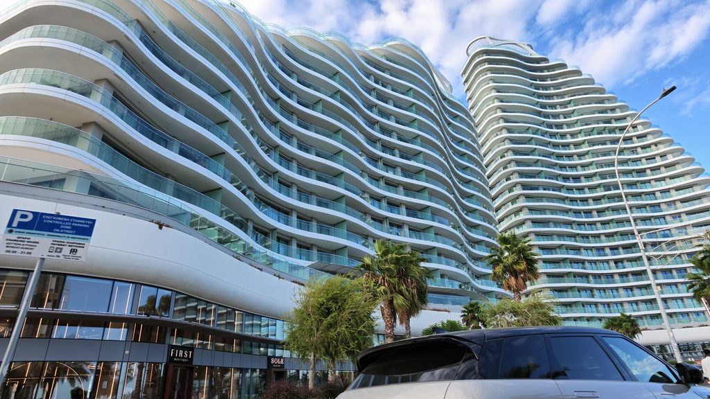 Two tall residential towers with wave-shaped white balconies and glass rails rise behind palm trees on the coast road east of Limassol.