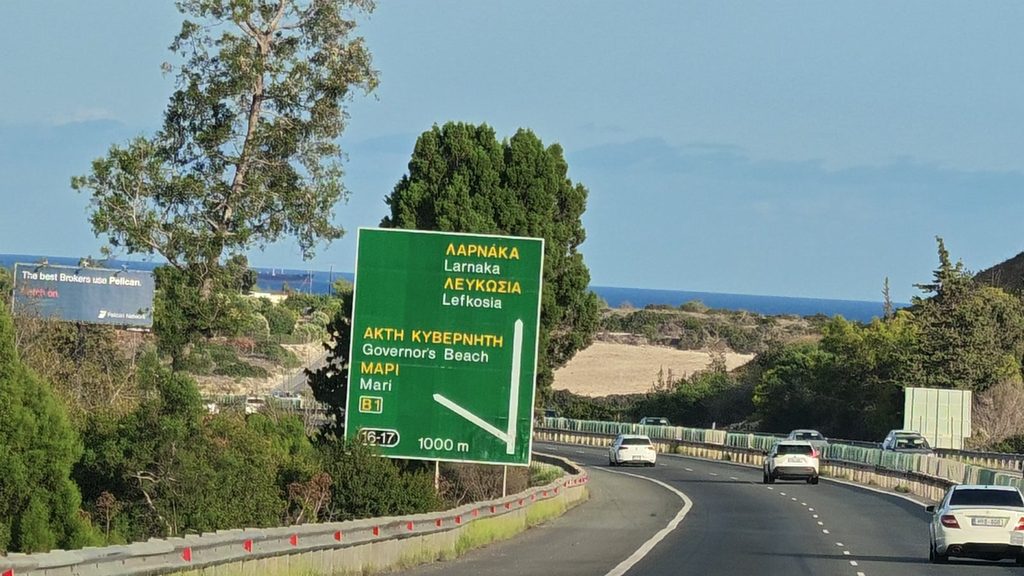 A green motorway sign on the motorway east of Limassol pointing straight on to Larnaka and Lefkosia and to the Governor's Beach and Mari exit in 1000 metres, with the sea beyond.