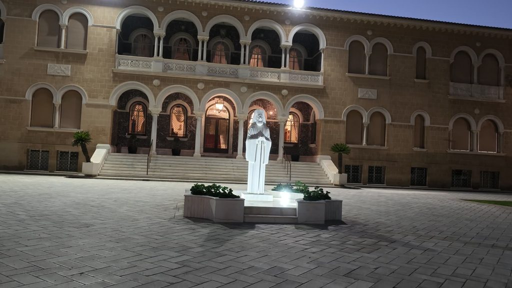 A two-storey arcaded building in old Nicosia at blue hour, its arches lit from within, with a white statue on a lit plinth in the paved square in front.