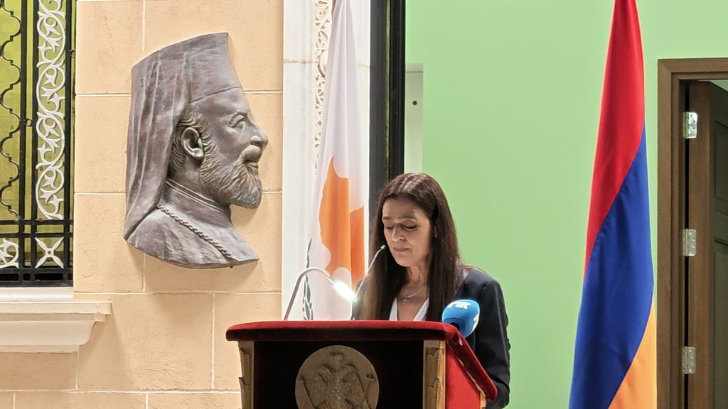 A woman with long dark hair reading at a wooden lectern, beside a bronze relief of Archbishop Makarios and between the flags of Cyprus and Armenia.