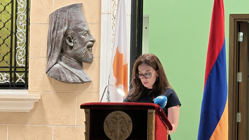 A woman in glasses reading at the same wooden lectern, with the bronze relief of Archbishop Makarios behind her and the Armenian flag at the right.