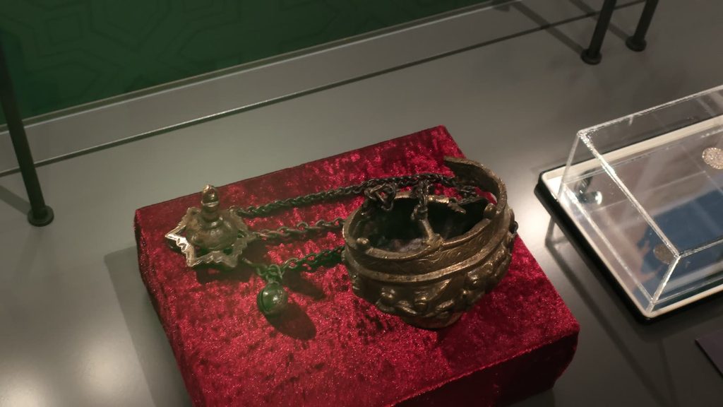 A bronze censer, an open bowl with figures in relief round its side, lying with its chains and their star-shaped top plate on a red velvet stand in a glass case.