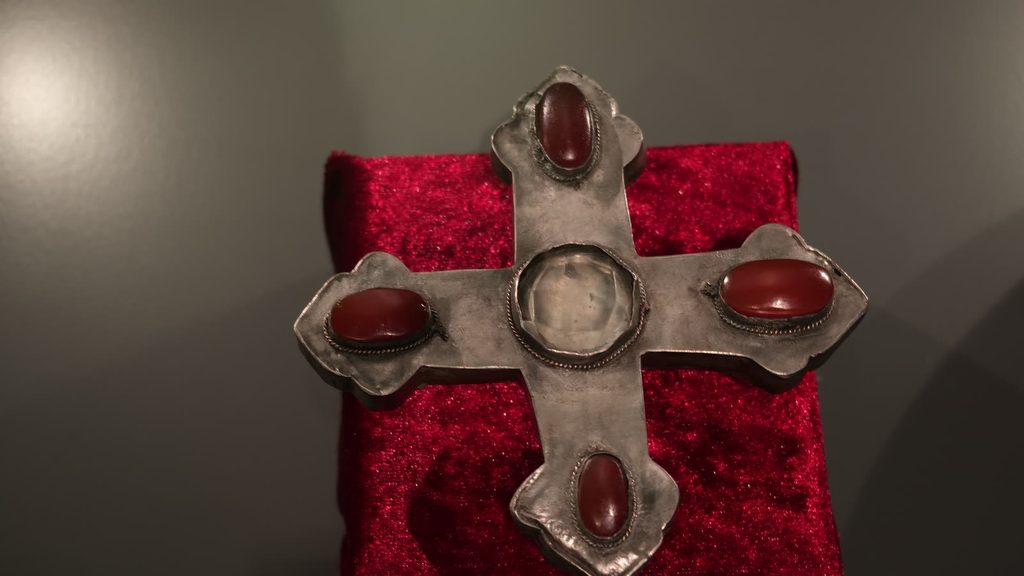A silver cross with flared arms, set with four oval red stones and a round clear stone at its centre, on red velvet.