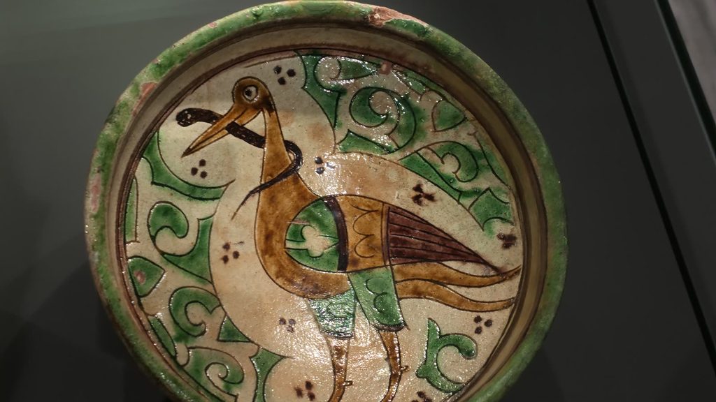 A glazed ceramic dish painted with a long-legged bird in brown and green among green leaf scrolls on a cream ground.
