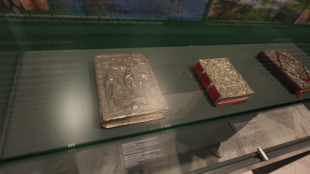 Three Gospel books in silver covers worked in relief, one with a crucifixion scene, lying in a glass case below a printed wall panel.