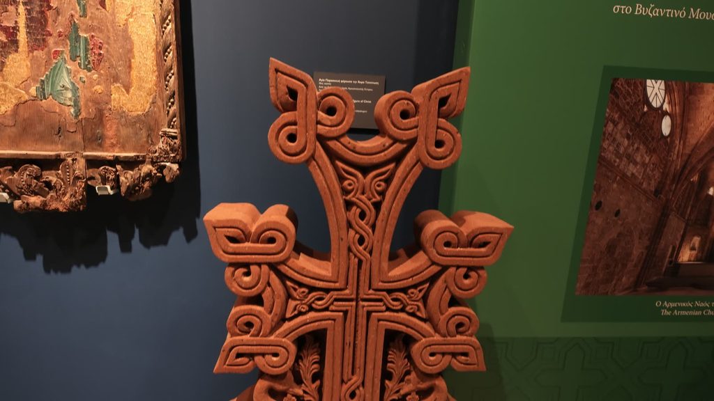 The top of a red stone khachkar carved with a cross of interlaced knotwork, against a dark blue wall and a green panel, beside the carved wooden frame of an old icon with worn gilding.