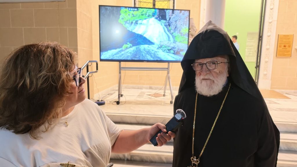 Araz, in a white top, holds a microphone to Archbishop Gomidas Ohanian, white-bearded in a black hood and robe with a gold chain and crowned pendant, in the museum's marble hall.